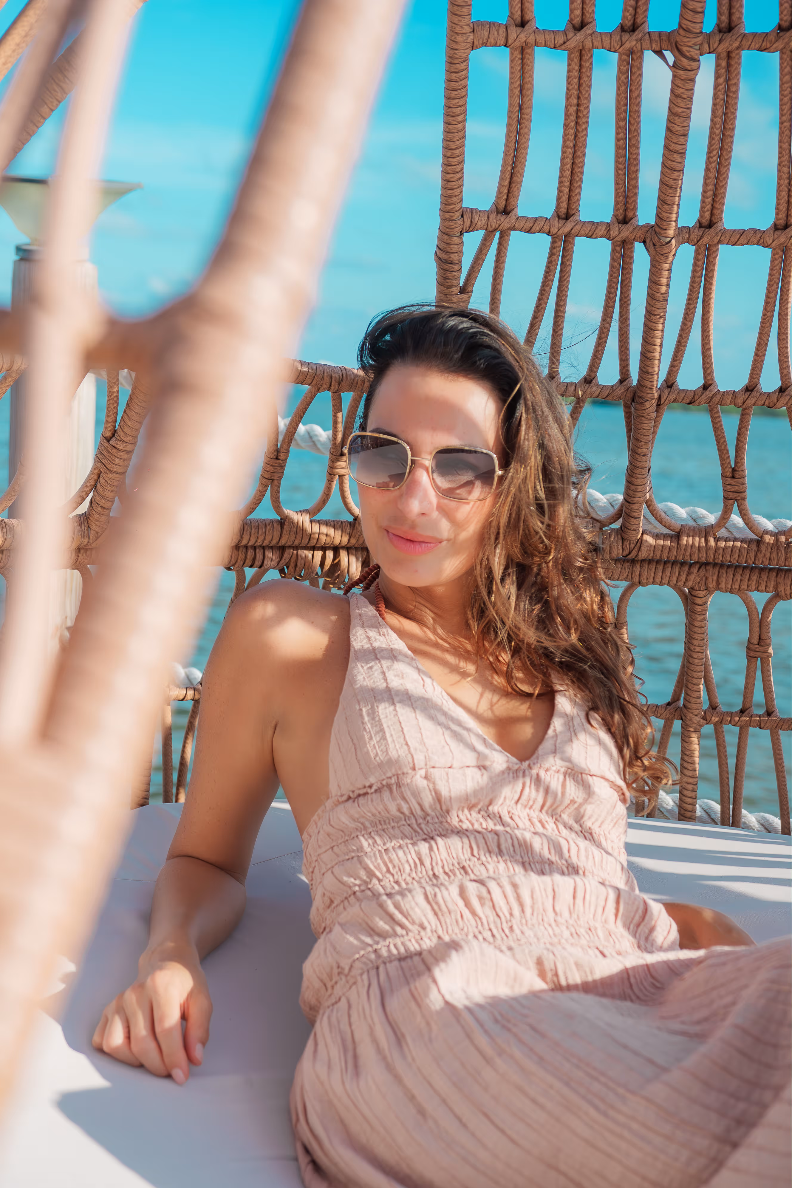 Woman wearing sunglasses and a sleeveless dress relaxing in a woven hammock chair by the water.