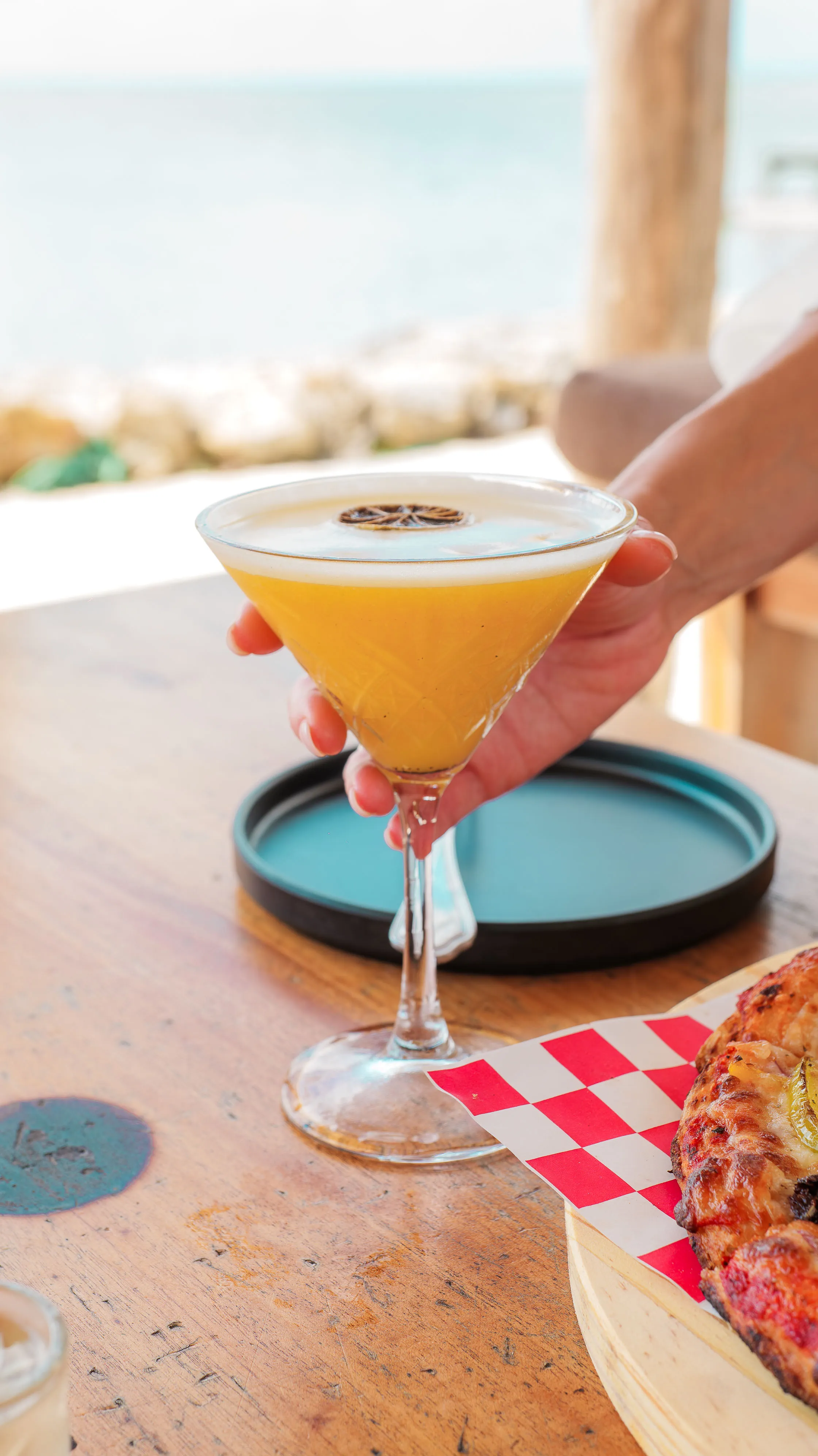 Hand holding a yellow cocktail in a martini glass with a dried citrus garnish on a wooden table next to a pizza.