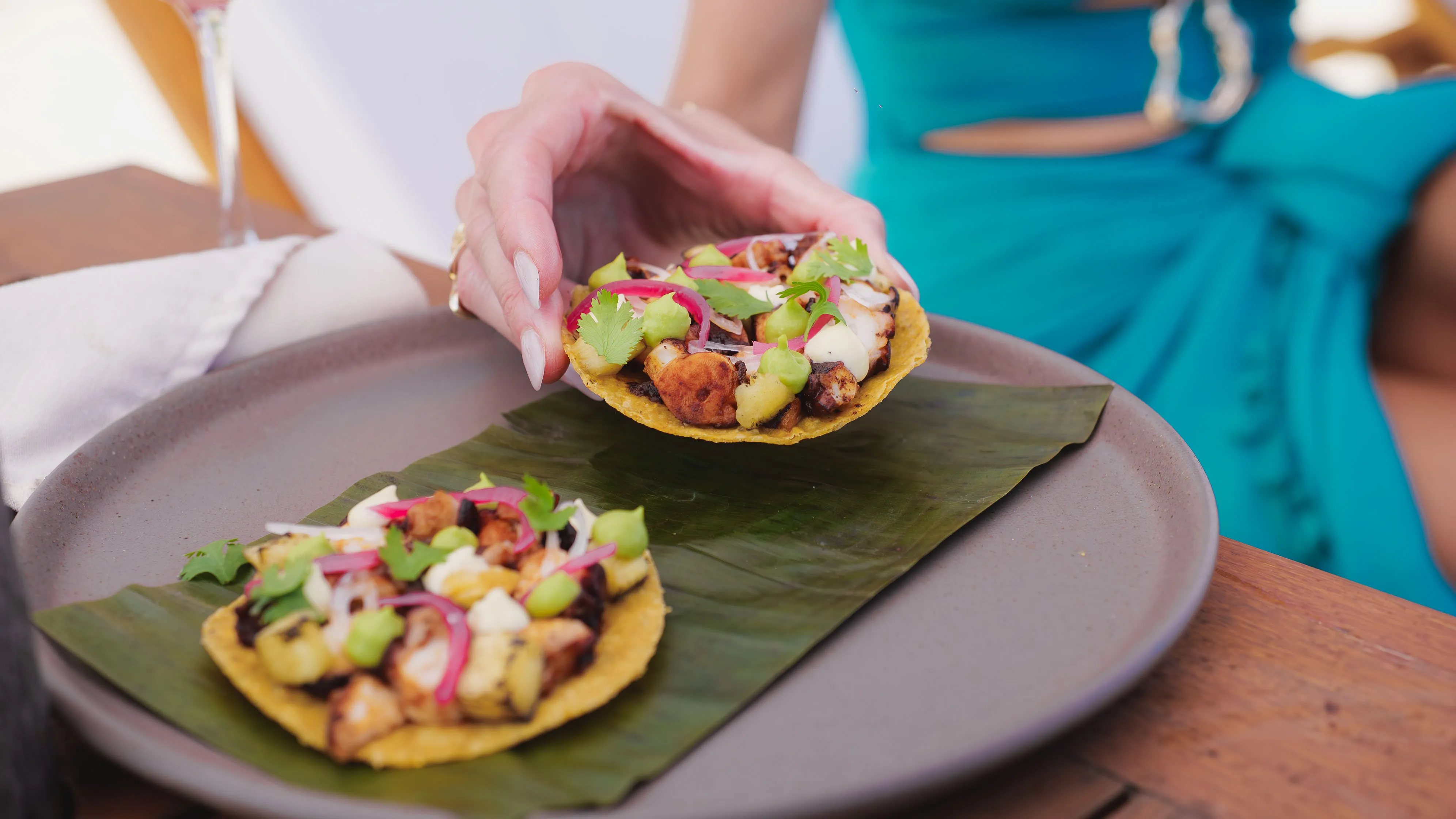 Two tostadas topped with grilled octopus, avocado, pickled onions, and cilantro on a banana leaf in a grey plate, one being held by a person in a teal outfit.