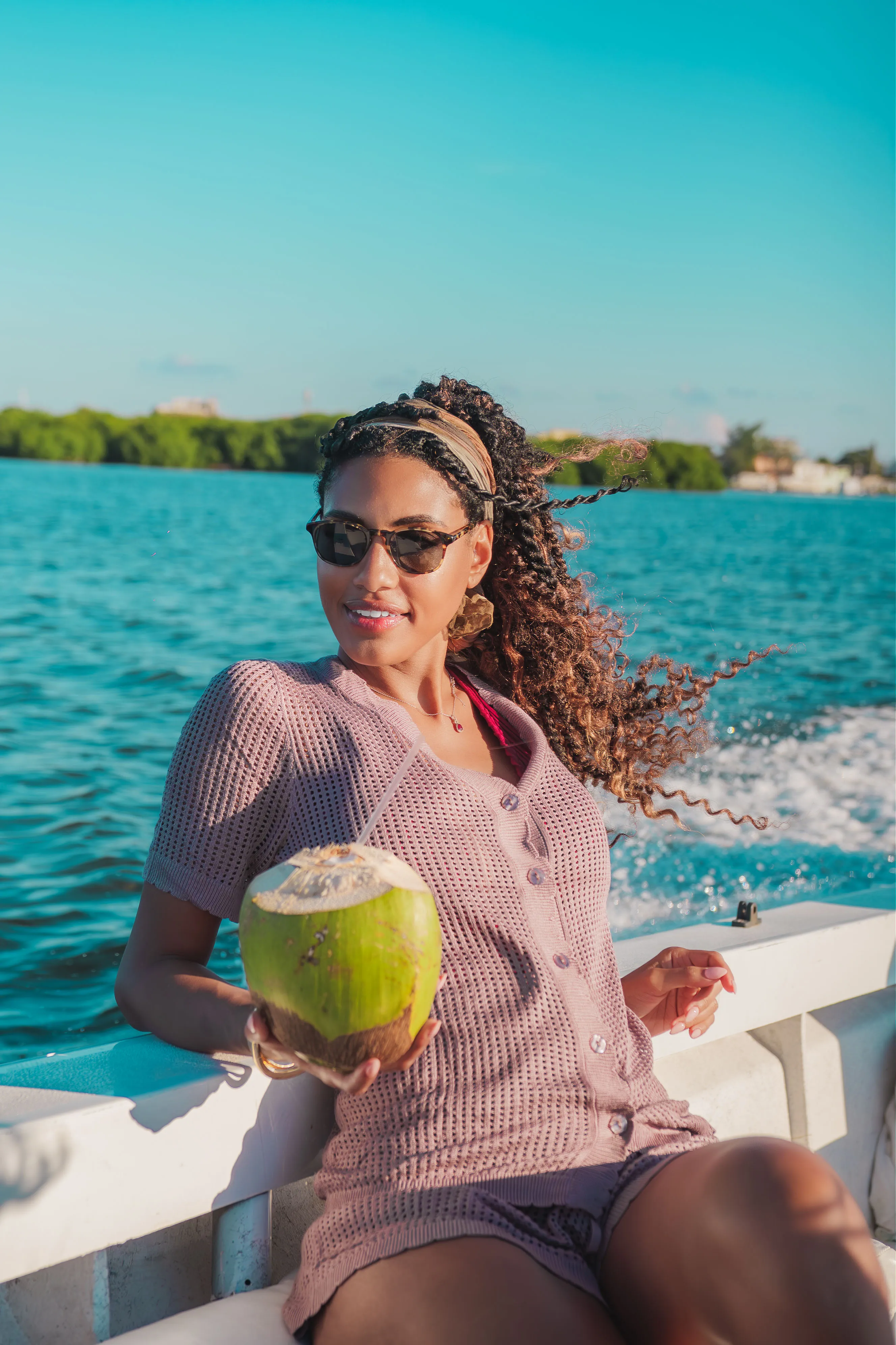 Woman with curly hair and sunglasses sitting on a boat holding a green coconut with a straw, with blue water and trees in the background.