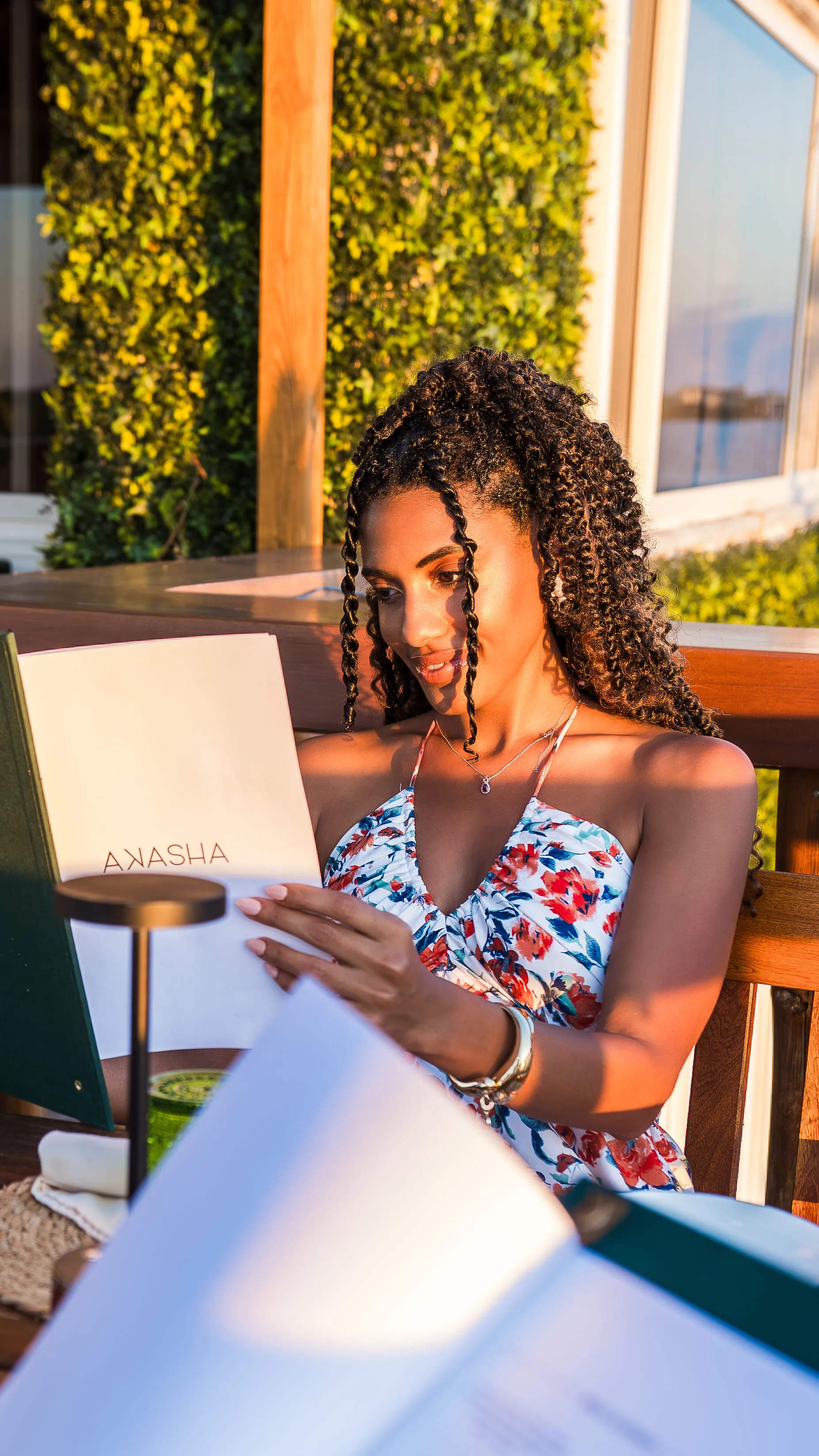 Woman with curly hair in a floral dress reading a menu outdoors at sunset.