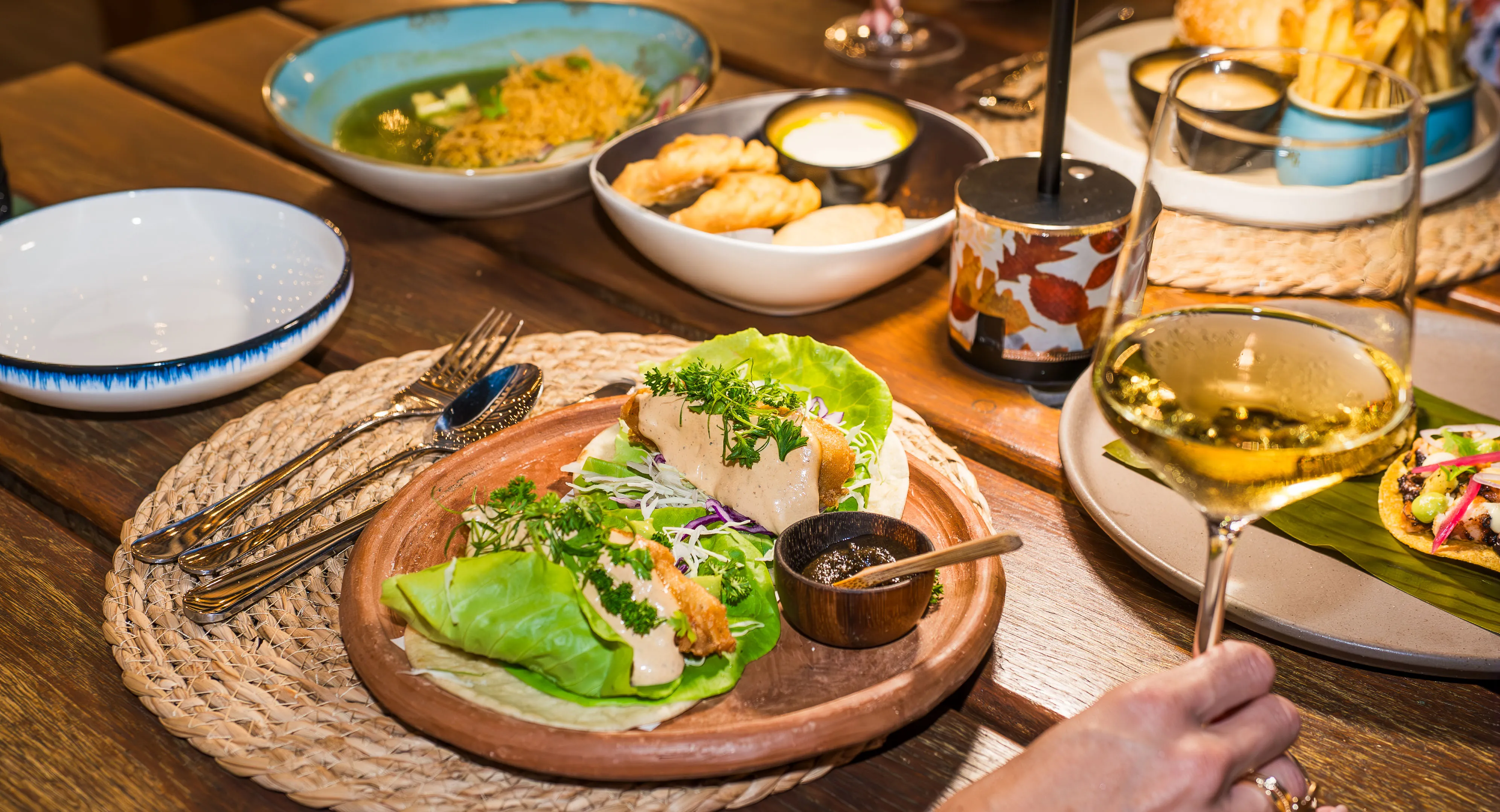 Wooden plate with two lettuce-wrapped tacos topped with creamy sauce and fresh herbs, with a small bowl of sauce, silverware, and a hand holding a glass of white wine on a wooden table.