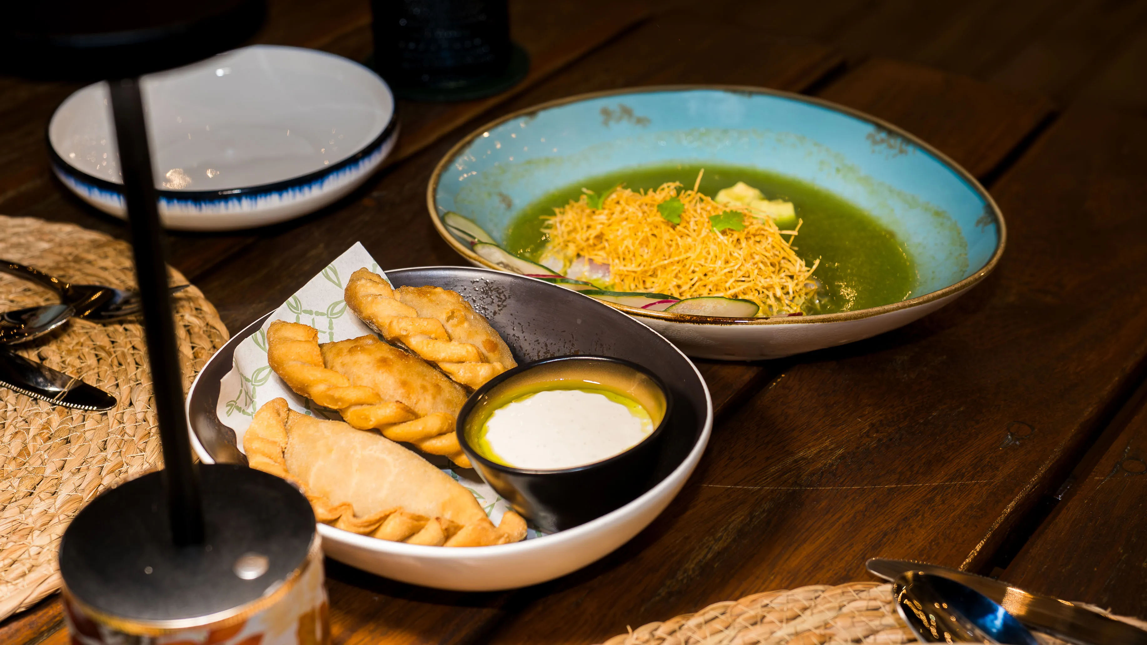 A bowl with three fried empanadas and a small dish of creamy dipping sauce, next to a blue bowl with green soup topped with shredded crispy garnish.