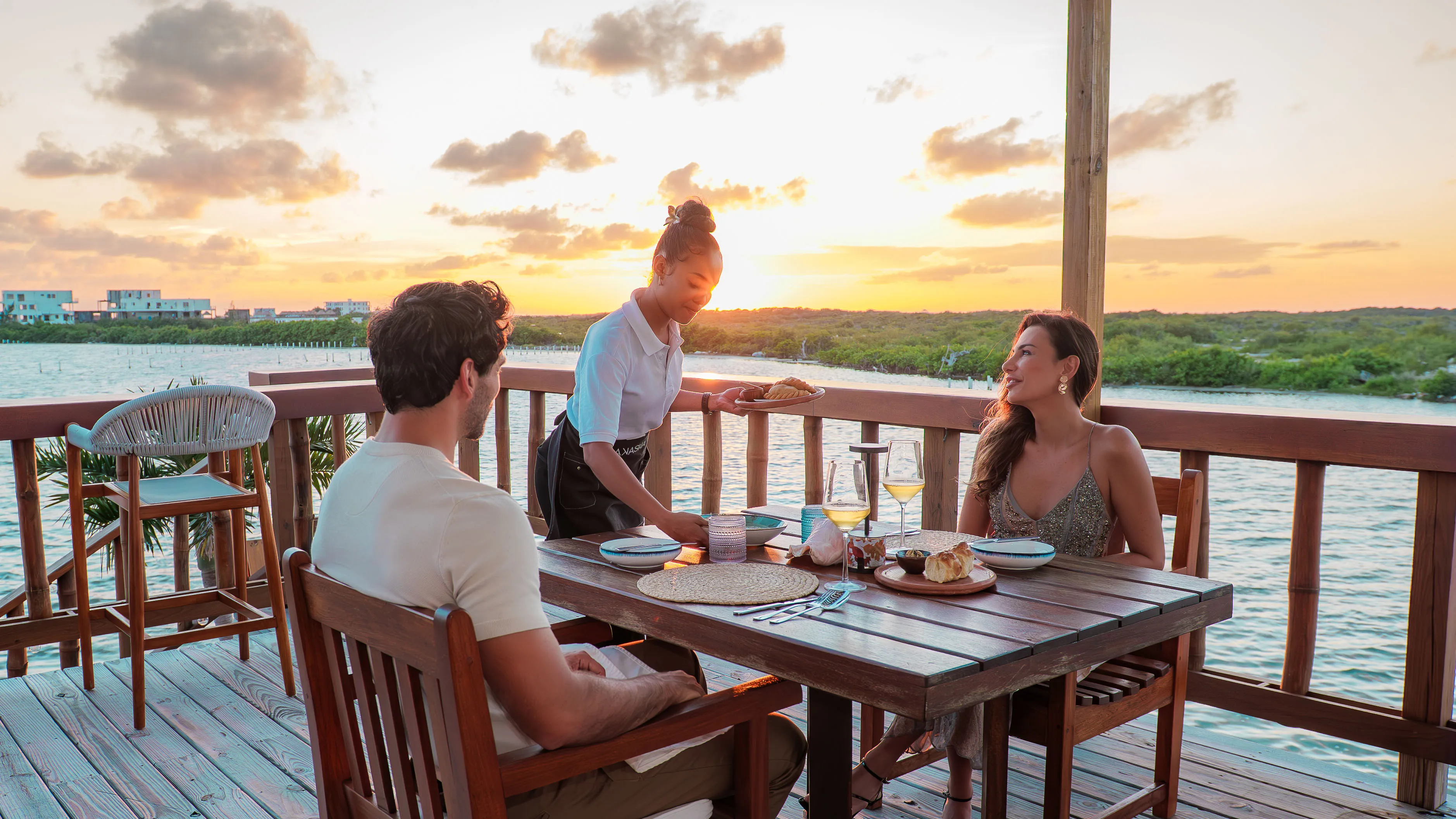 A couple dining outdoors at sunset by the water while a waitress serves them food on a wooden deck.