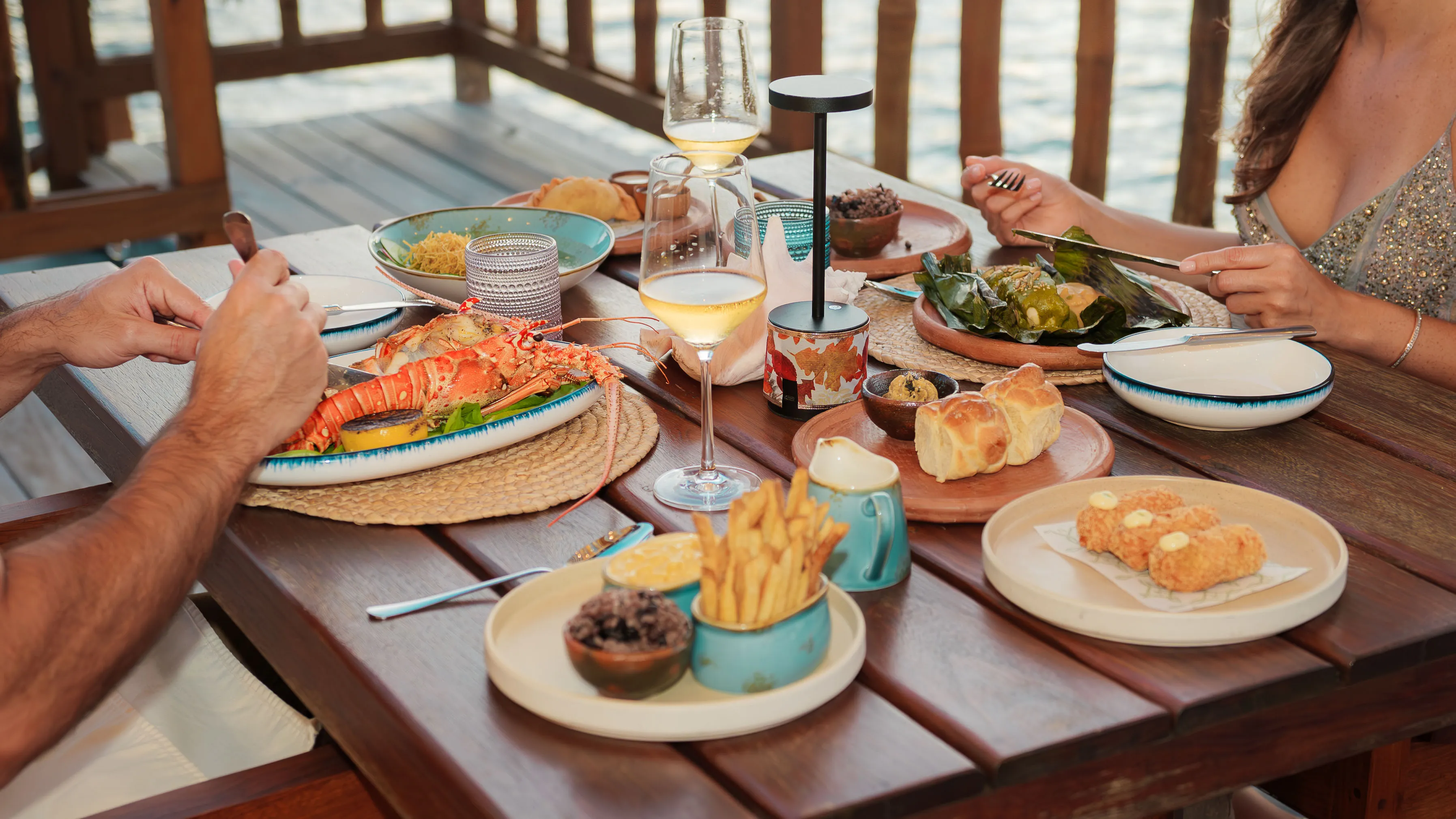Couple dining outdoors at wooden table with lobster, fries, bread, and wine glasses.