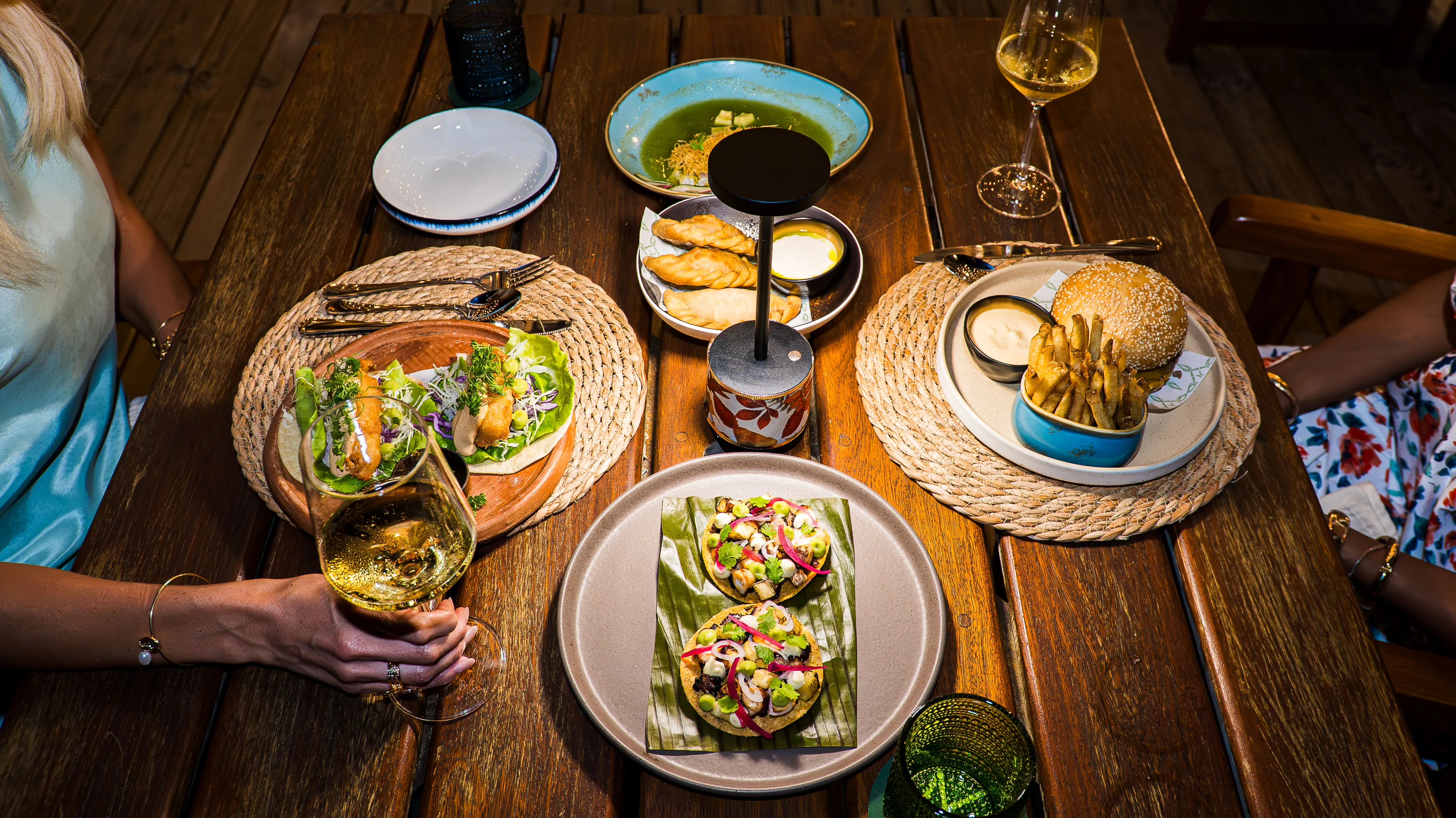 Table with a variety of dishes including small tacos, empanadas, a burger with fries, and two glasses of white wine, with two people seated at the table.