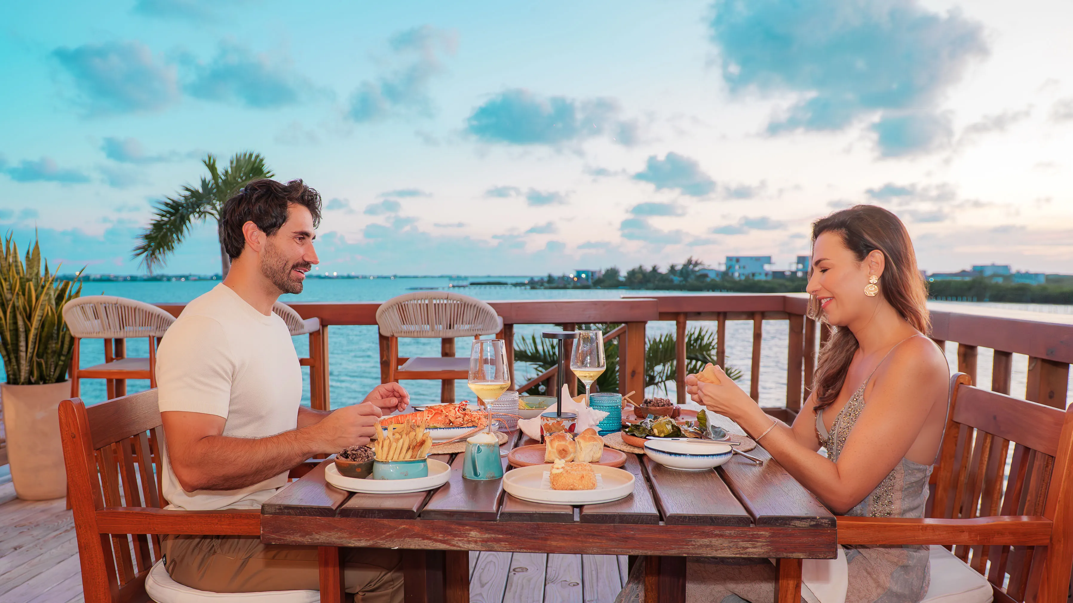 Man and woman enjoying a seafood dinner at a wooden table on a lakeside deck during sunset.