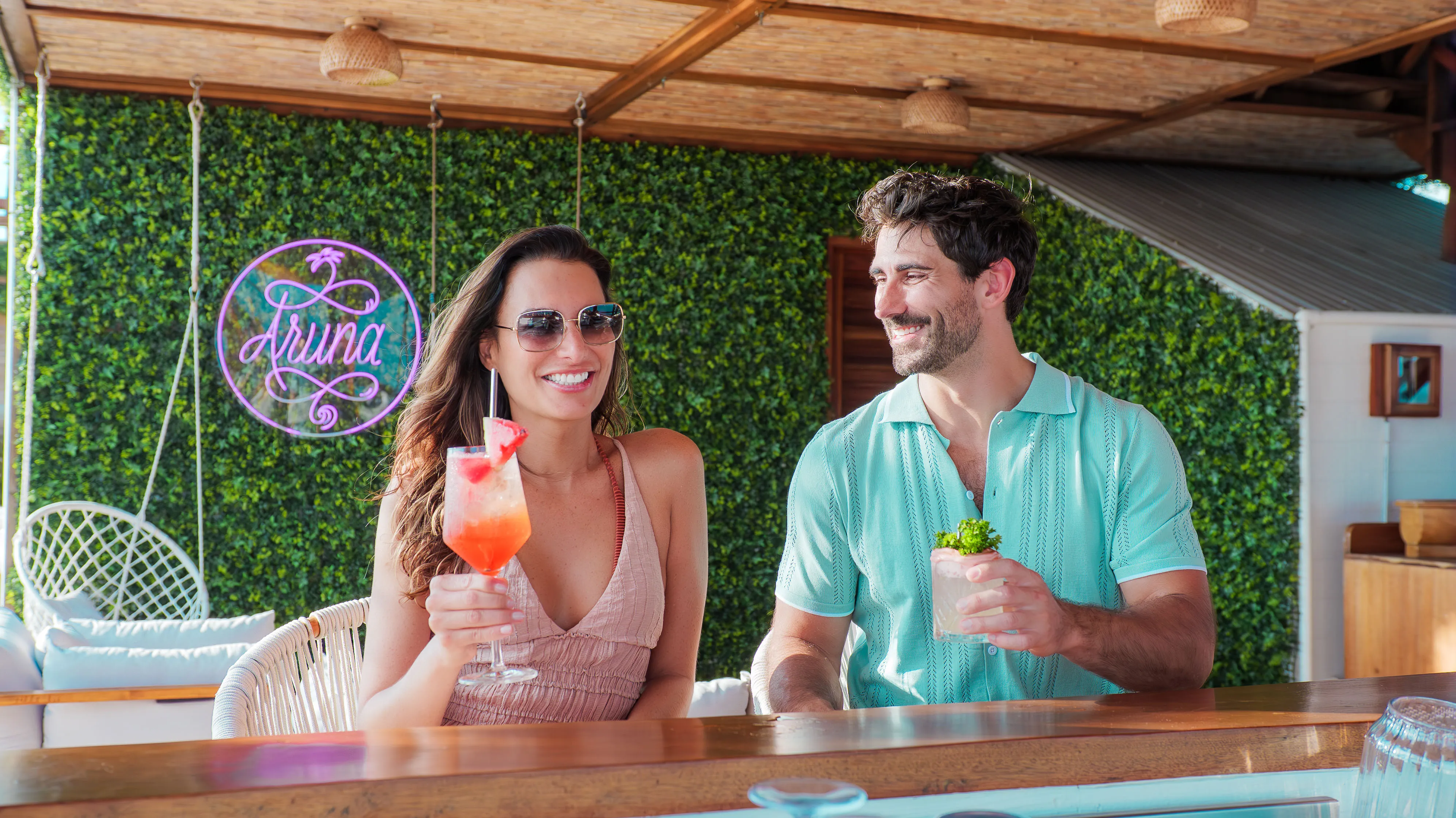 Smiling couple sitting at a bar with tropical cocktails, green foliage wall, and Aruna neon sign in the background.