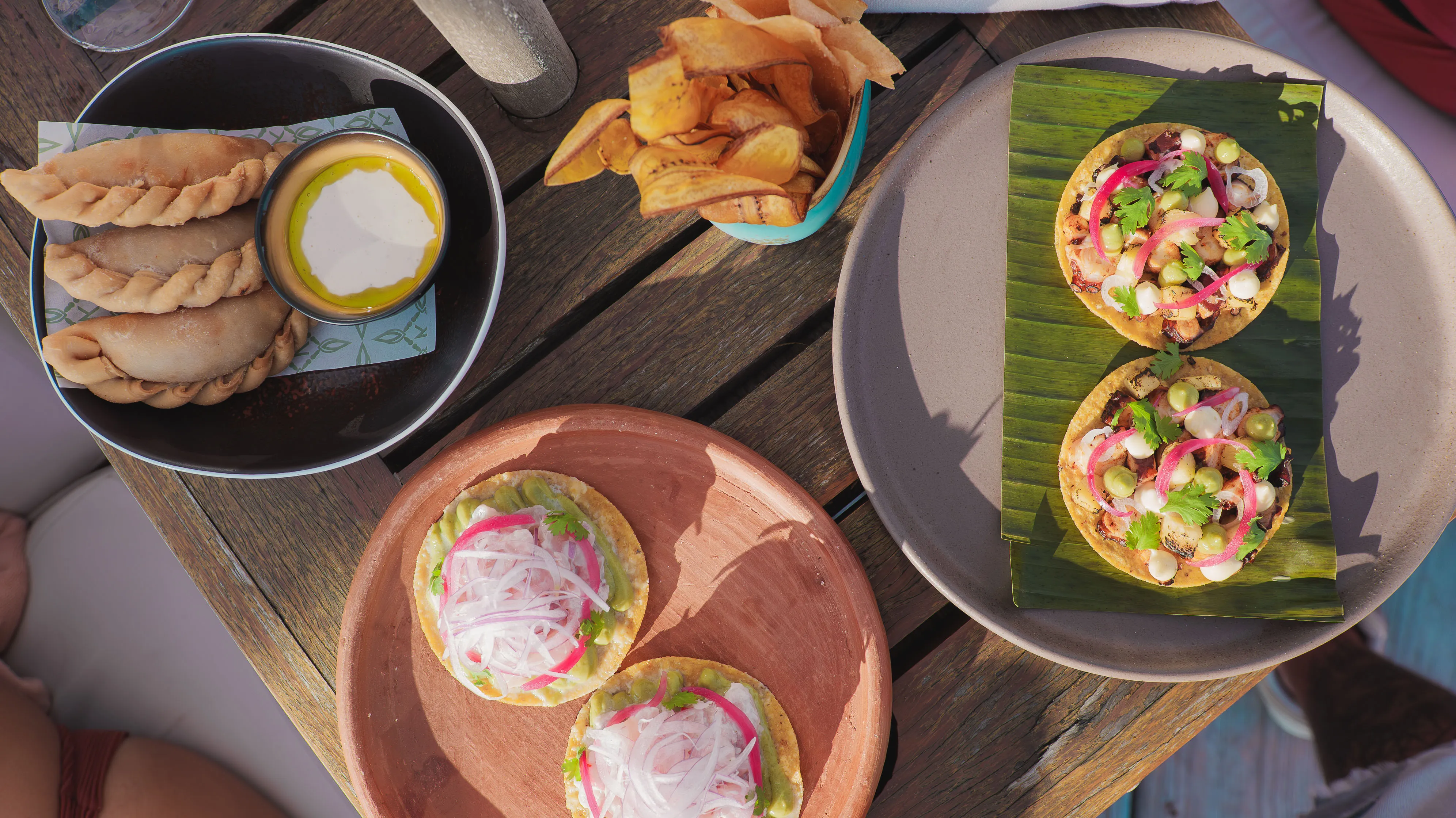 Table with plates of empanadas, plantain chips, and tostadas topped with avocado, pickled onions, and herbs.