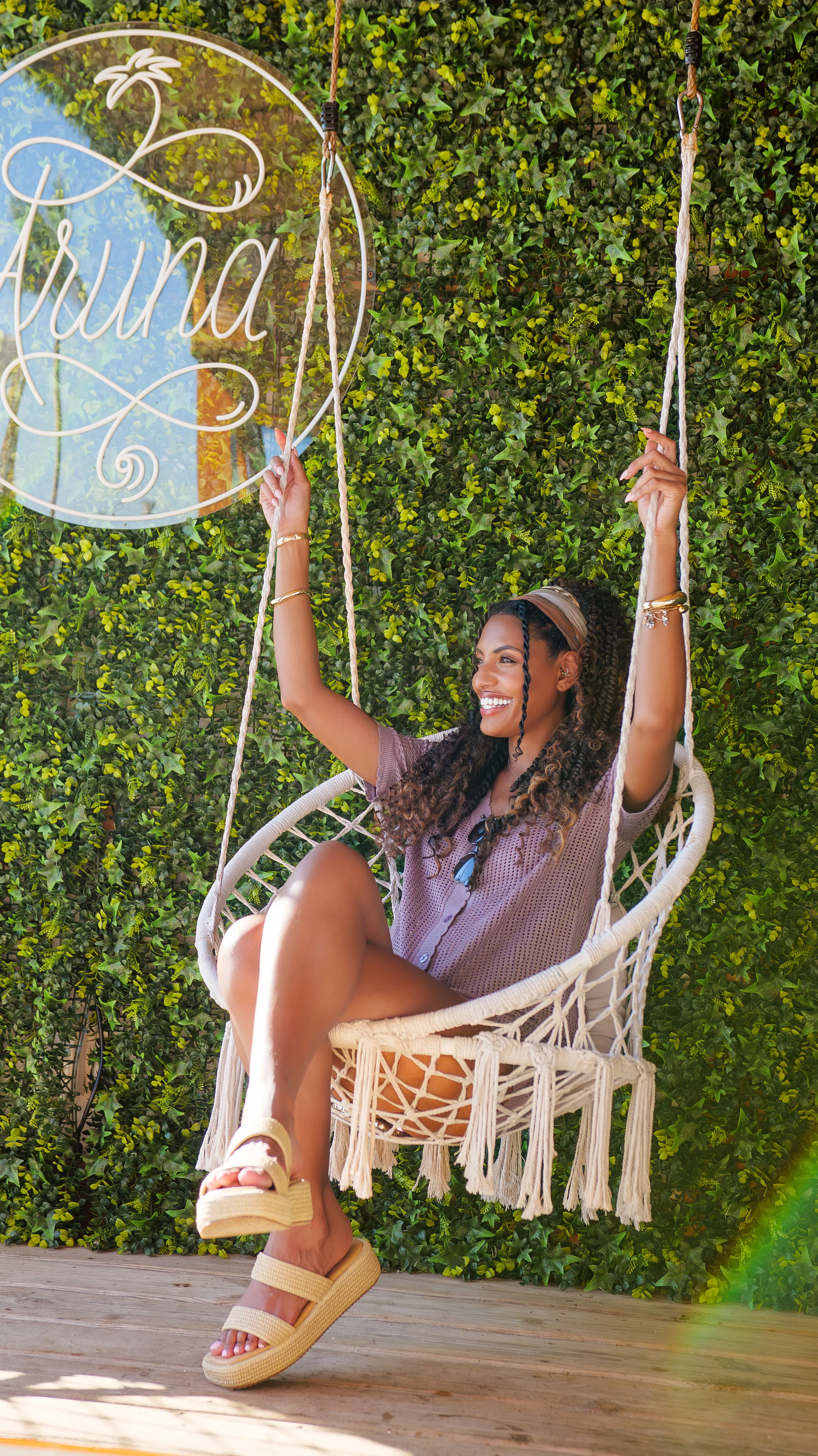 Smiling woman with curly hair sitting on a white macrame swing chair in front of a green leafy wall with a 'Parina' sign.