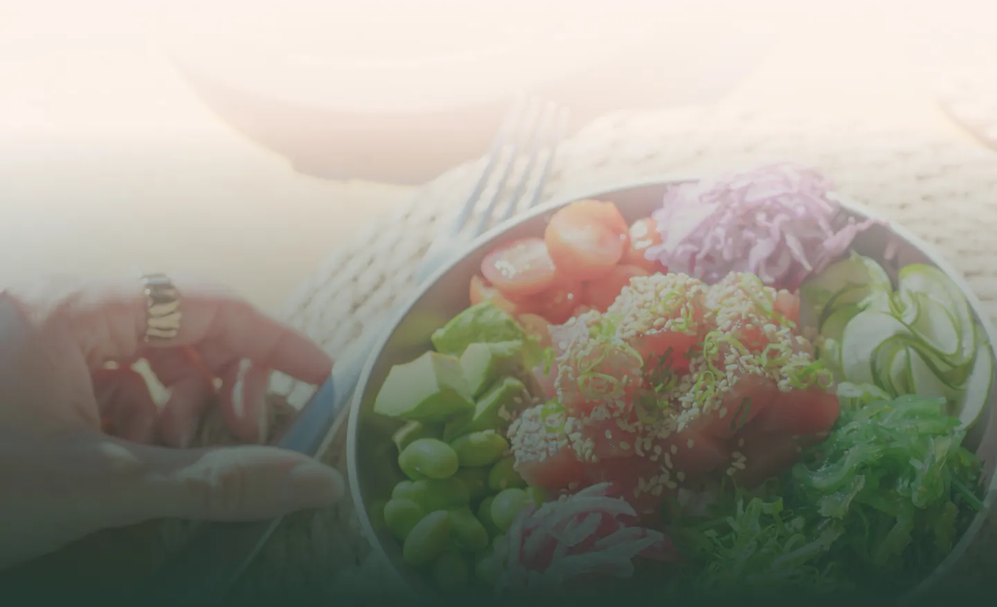 Hand holding a fork next to a bowl of fresh poke salad with tuna, avocado, edamame, cucumber, seaweed, cherry tomatoes, and shredded cabbage.