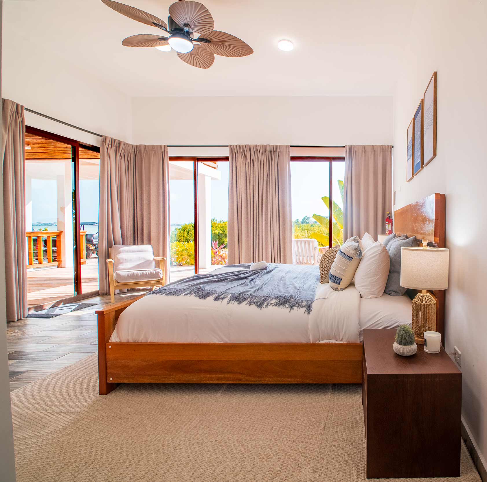 Bright bedroom with wooden bed, neutral bedding and pillows, nightstand with lamp and plant, ceiling fan, and large glass doors opening to a sunlit patio with greenery.