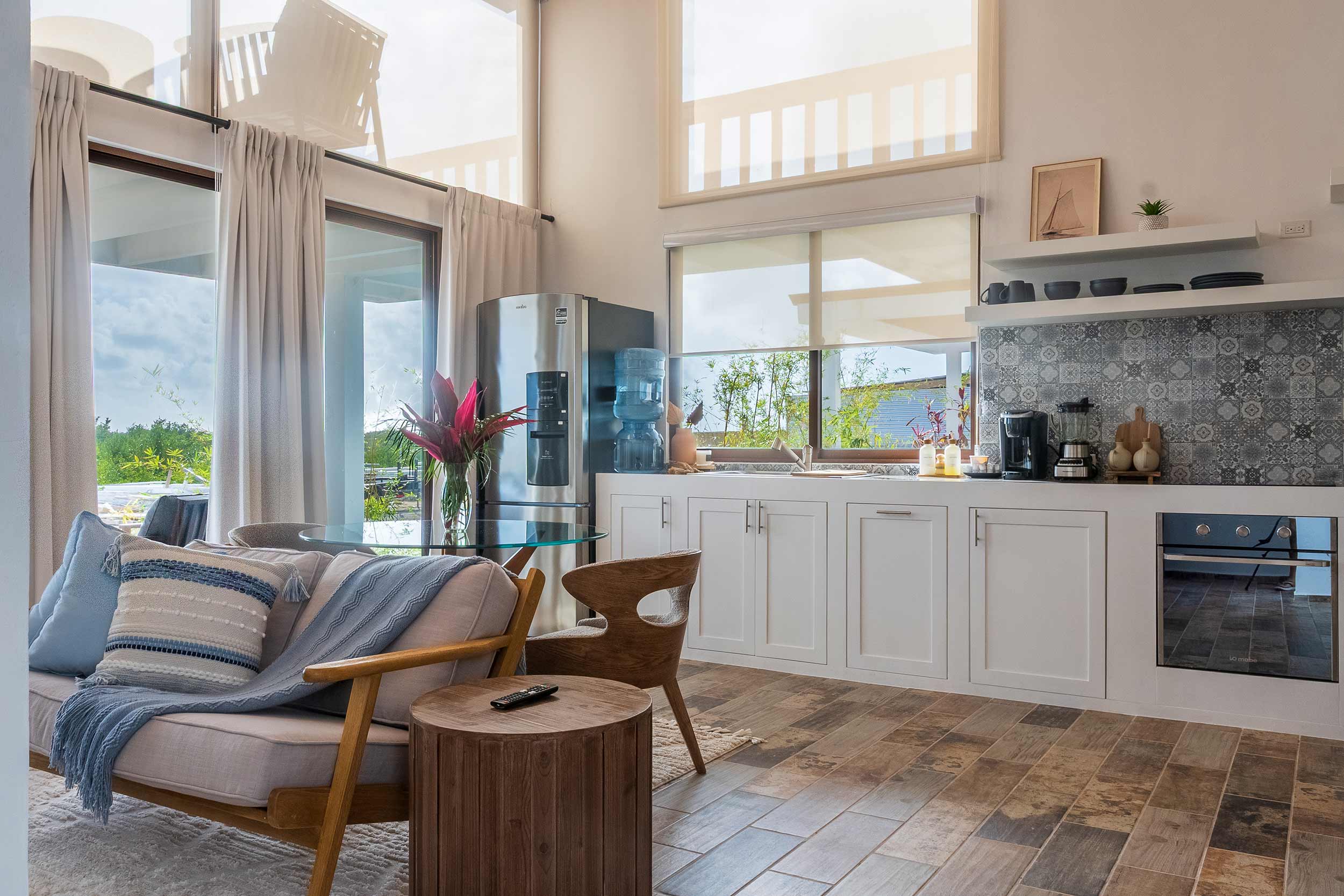Modern kitchen with white cabinets, patterned gray backsplash, stainless steel refrigerator, water dispenser, glass dining table with wooden chairs, and a cozy beige sofa with blue cushions and a throw blanket.