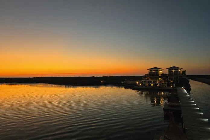 Calm water at sunset with an orange sky and two lit buildings connected by a pier on the right.