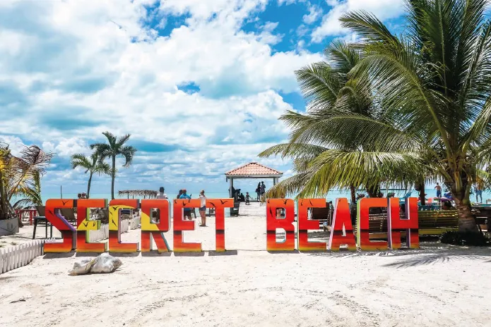 Large colorful letters spelling 'SECRET BEACH' on a white sandy beach with palm trees and a partly cloudy sky.