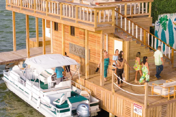 People walking on a wooden dock attached to a two-story wooden building labeled ‘AKASH’ with a white pontoon boat moored nearby.