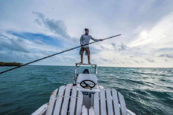 Man balancing on a boat while propelling it with a long pole over open water.