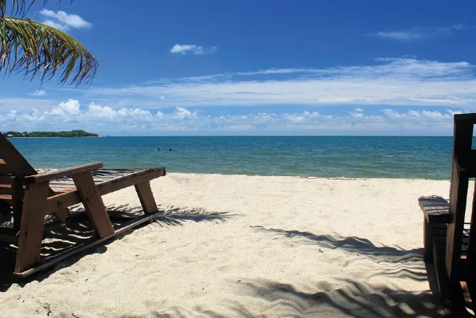 Wooden lounge chairs on a sandy beach with a calm sea under a blue sky with scattered clouds.