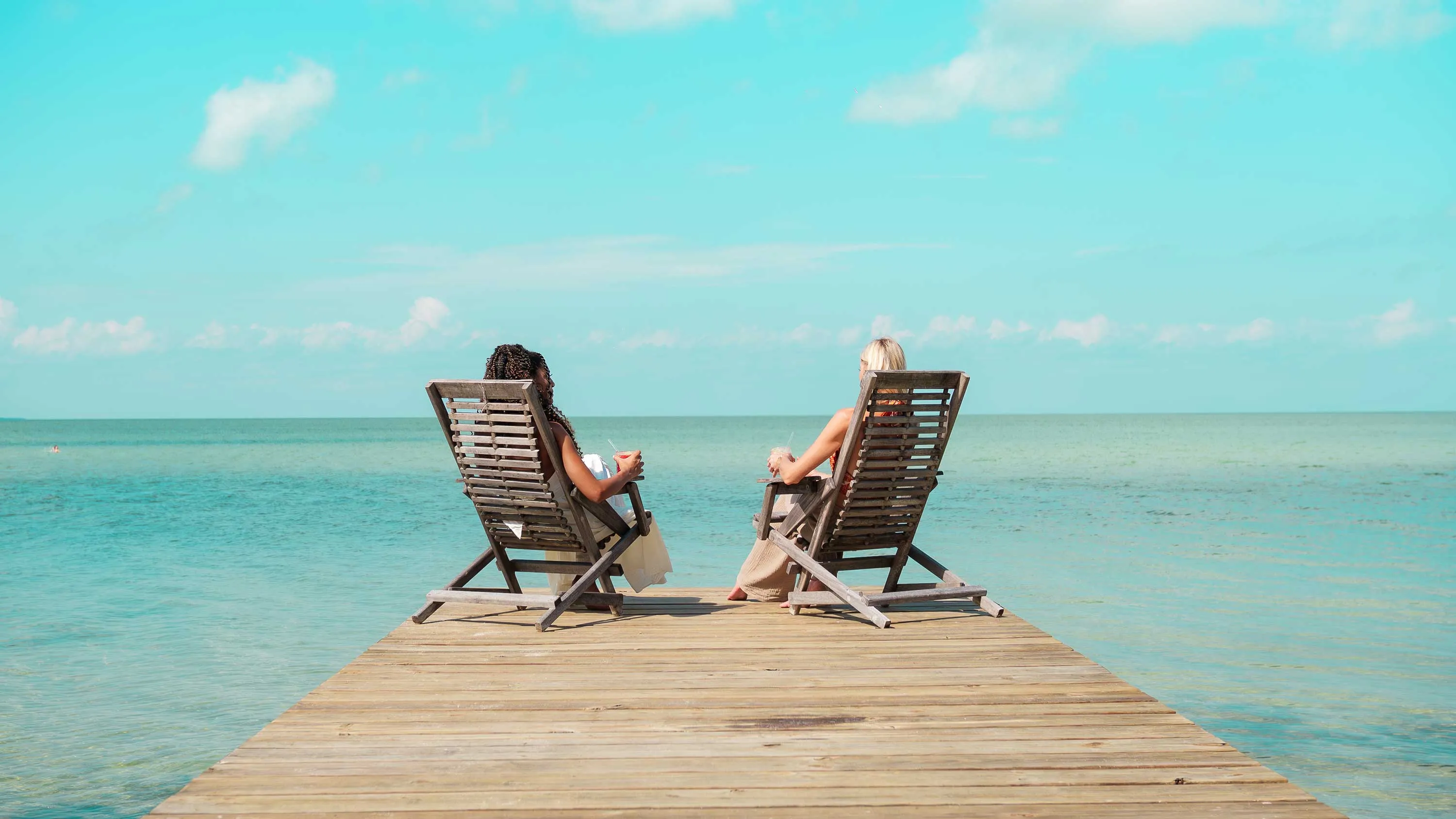 Two women sitting on wooden loungers on a jetty overlooking calm turquoise ocean under a partly cloudy blue sky.