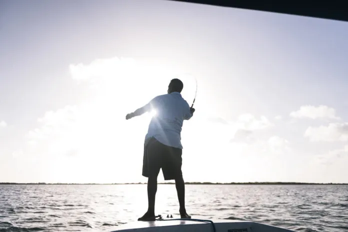 Person fishing from a boat at sunset, silhouetted against the sky over calm water.

