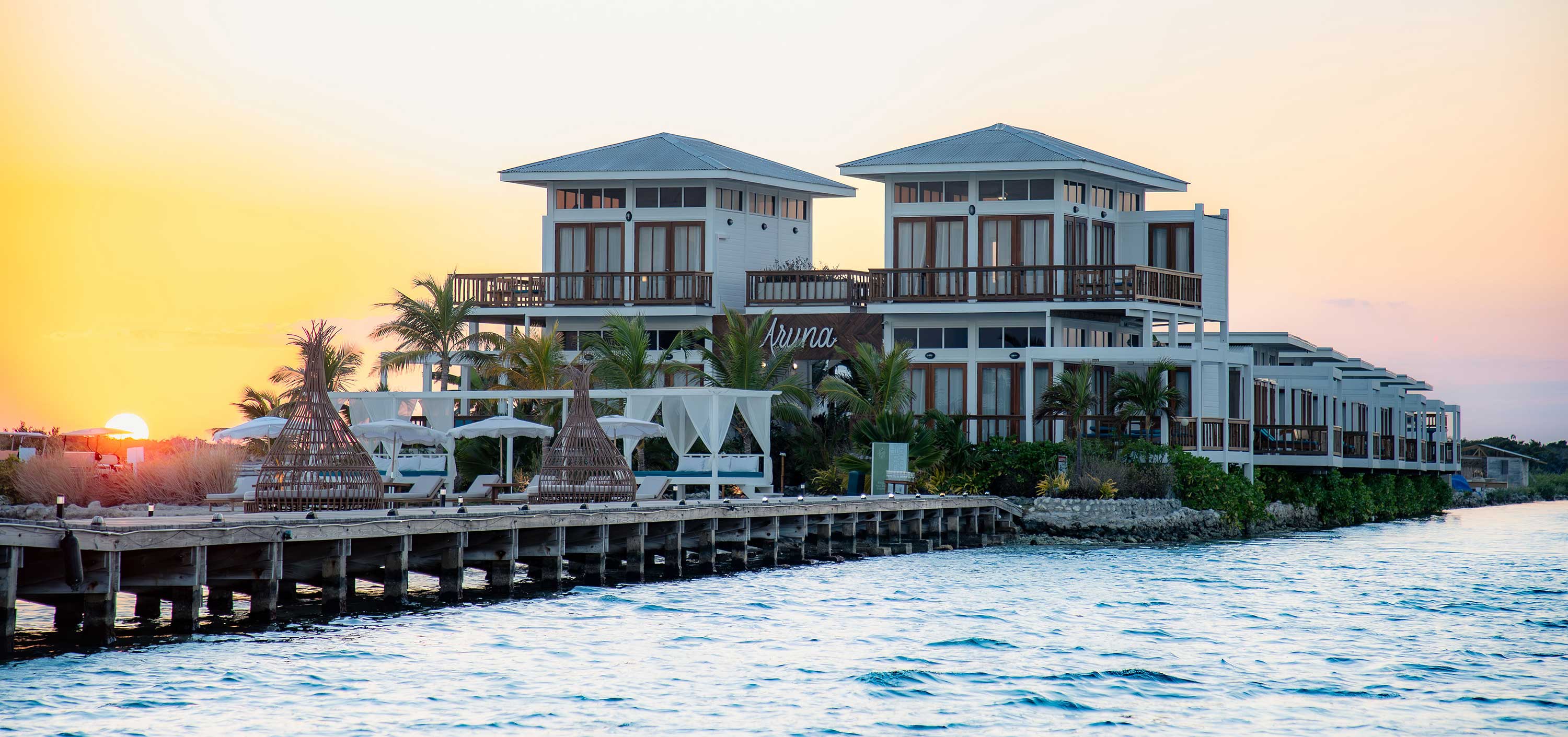 Modern white beachfront resort with palm trees, sun loungers, and wicker cabanas at sunset.