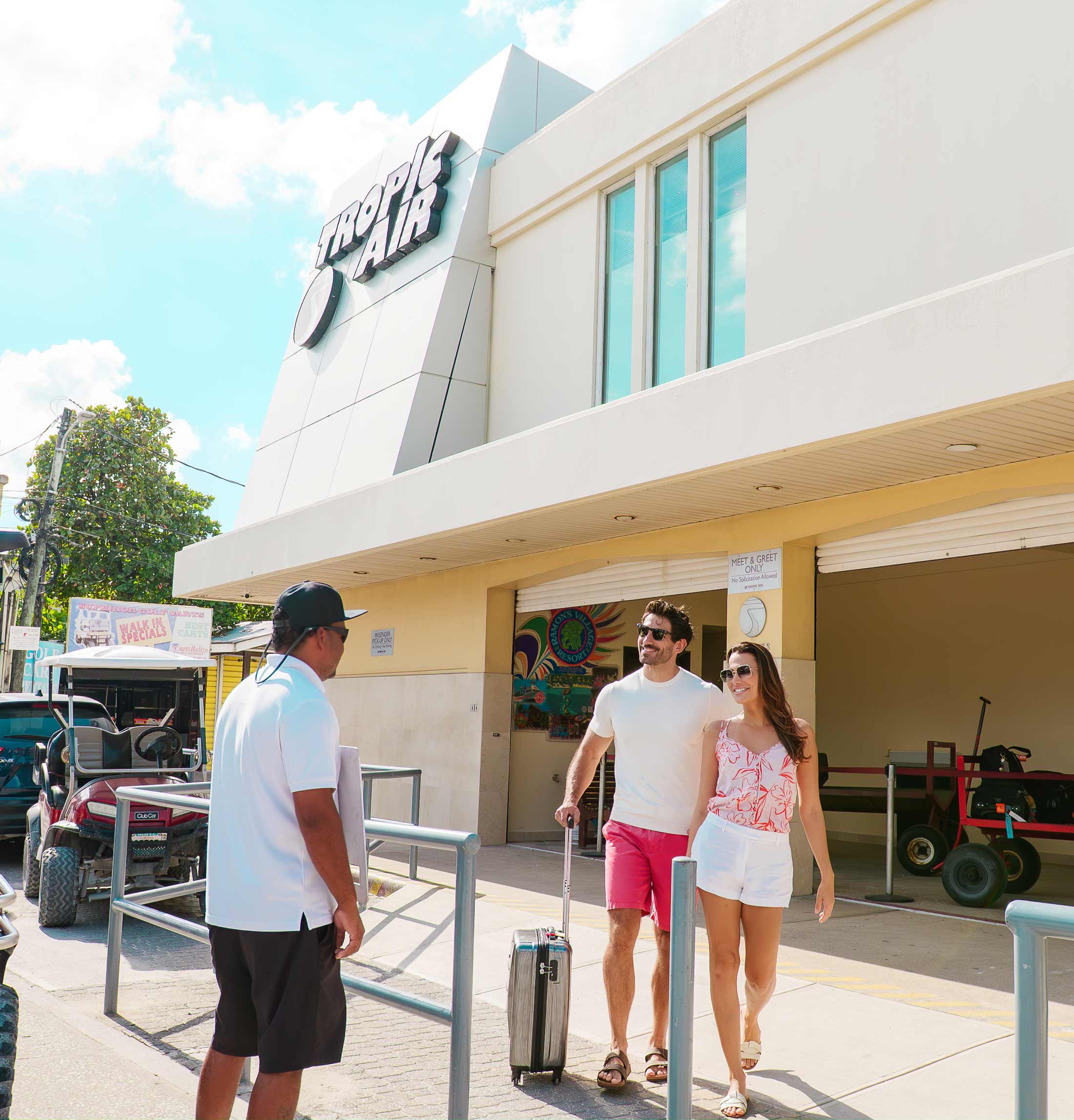 Couple with suitcase being greeted by a man outside Tropic Air building on a sunny day.