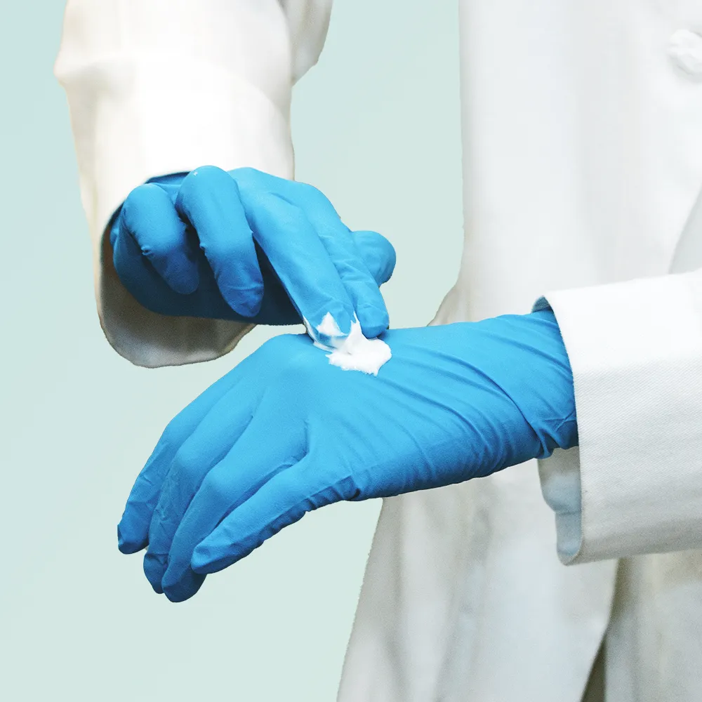 Person wearing blue medical gloves and a white coat applying a small amount of white cream on the back of one gloved hand using the fingers of the other gloved hand.