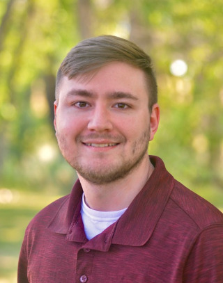 Young man with short brown hair and a light beard wearing a maroon collared shirt, smiling in an outdoor setting with blurred green foliage background.