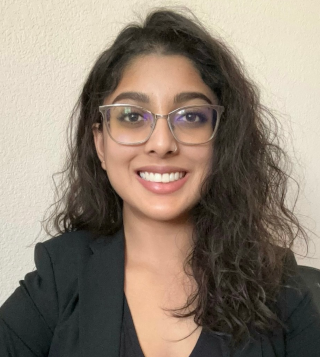 Smiling woman with curly dark hair and glasses wearing a black blazer in front of a plain wall.