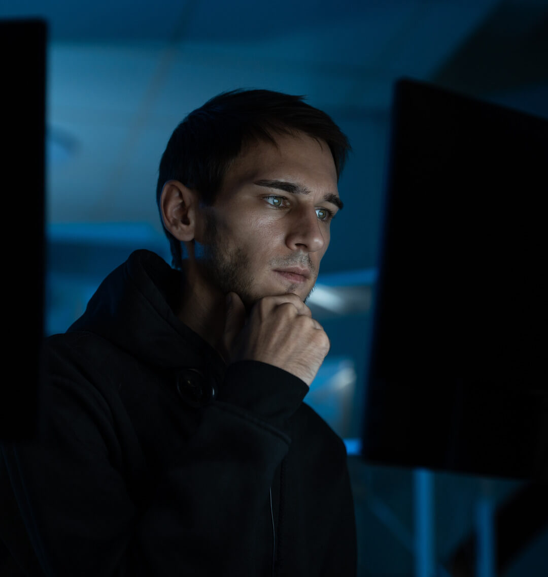 Young man in a dark hoodie thoughtfully looking at a computer screen in a dimly lit room.