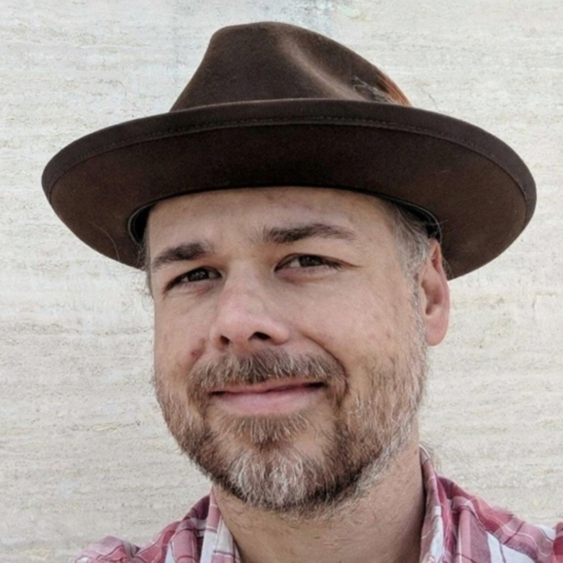 Portrait of a man with slicked-back hair wearing a blue collared shirt, outdoors with blurred greenery in the background.