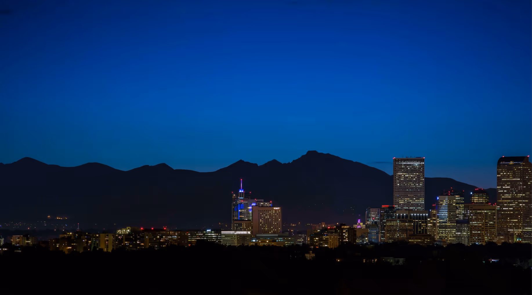 City skyline at dusk with illuminated buildings and dark mountain silhouettes under a deep blue sky.