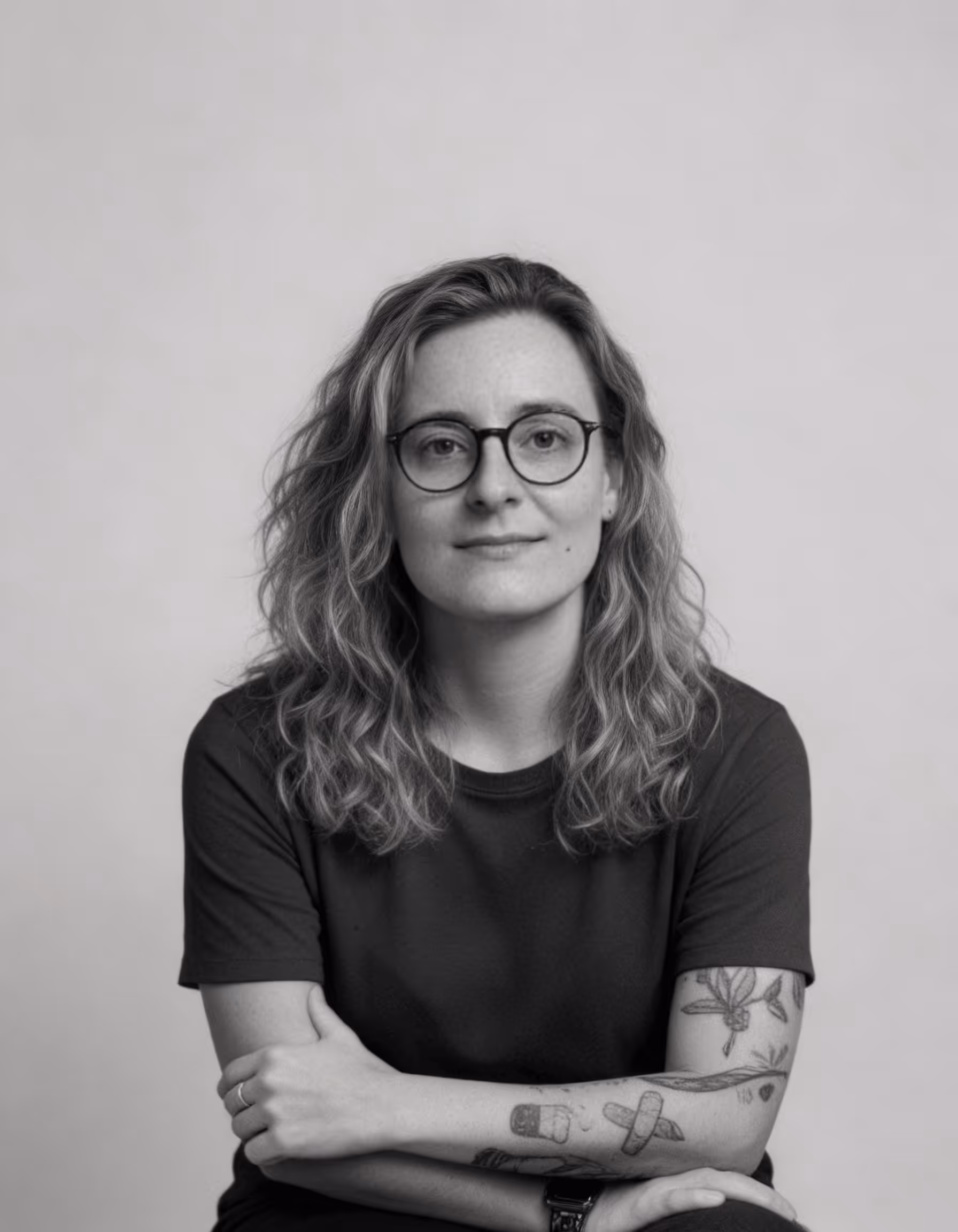 Retrato em preto e branco de mulher com cabelo ondulado, óculos e tatuagens no braço.