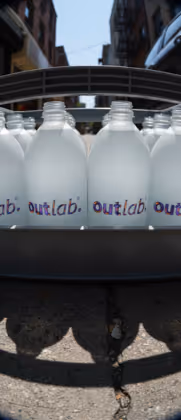 Garrafa branca com rótulo 'outlab.' em um carrinho sob luz do dia entre prédios.