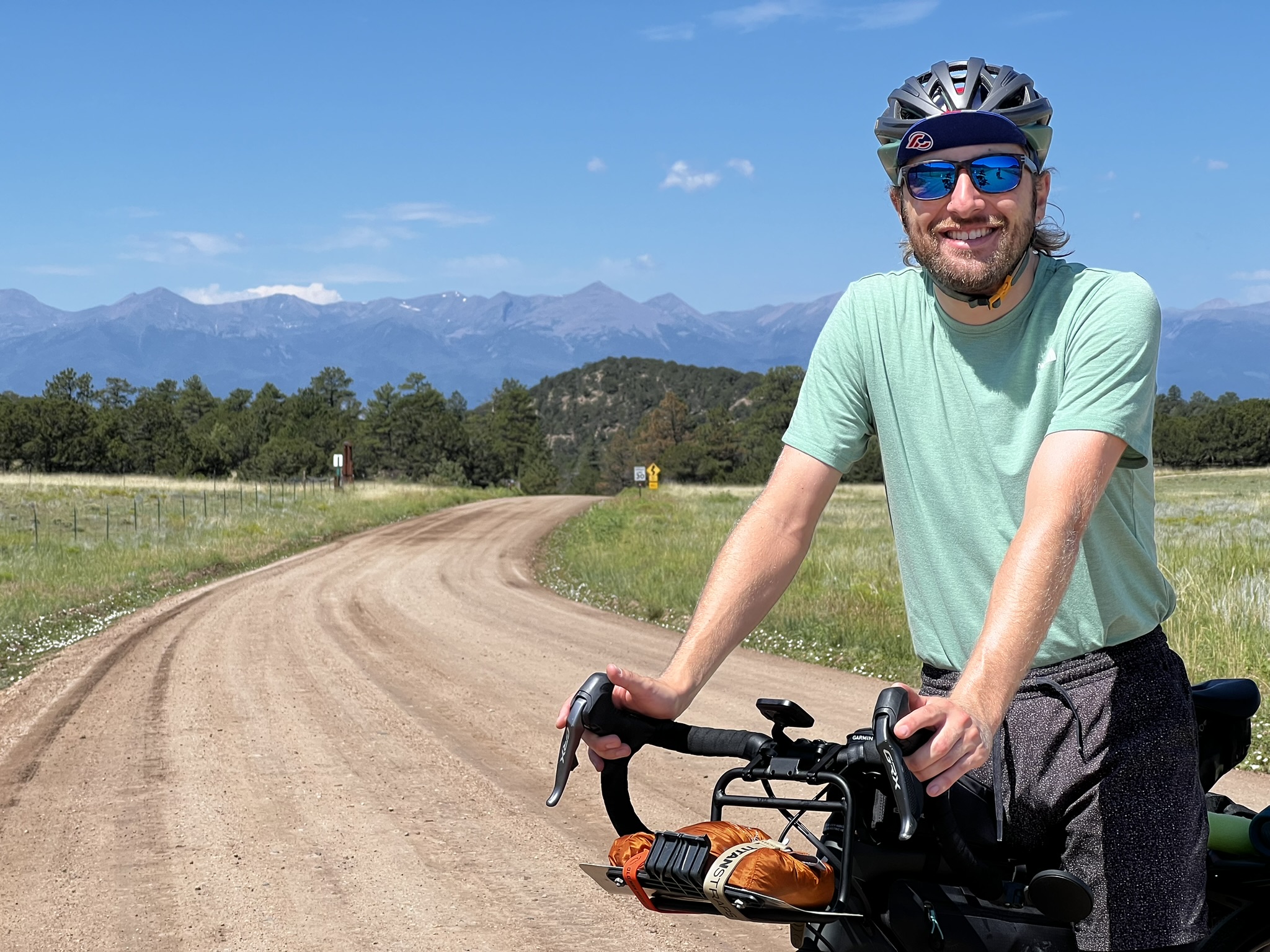 Dylan smiling beside a loaded bicycle on a dirt road with mountains behind him.