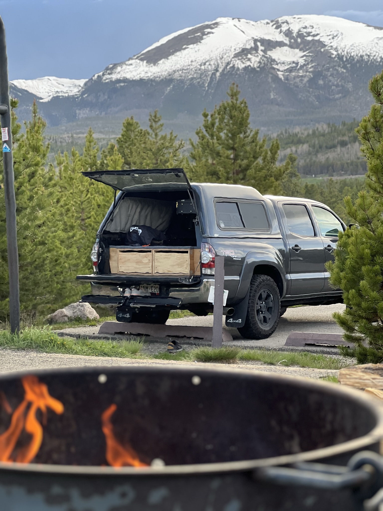 A truck camping setup beneath snowy mountains, with a campfire in the foreground.