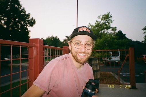 Dylan smiling outdoors in glasses, a cap, and a pink shirt.