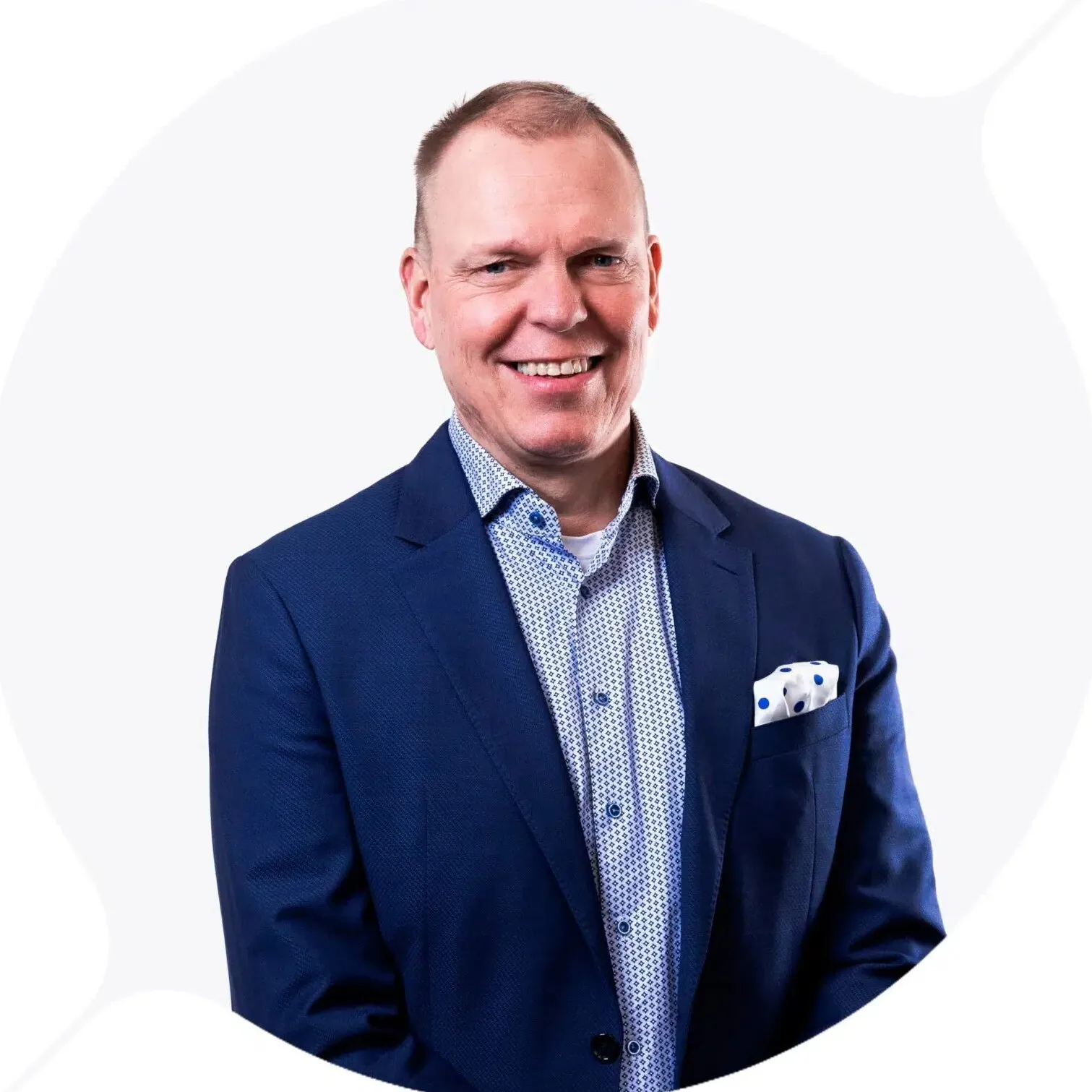 Hymyilevä, keski-ikäinen mies sinisessä puvussa ja valkoisessa paidassa, jossa on sinisiä kuvioita, valkoisella taustalla. Taskussa valkoinen nenäliina sinisillä pilkuilla.