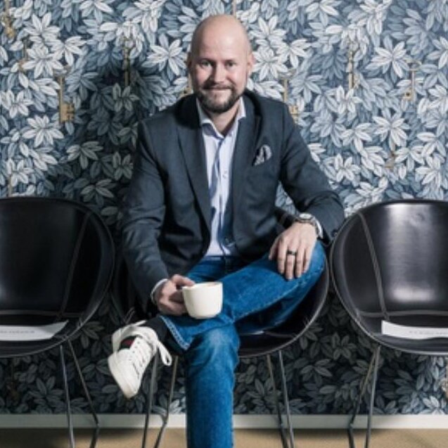 Keskellä istuva kalju mies, jolla on tumma pikkutakki, vaaleansininen paita, farkut ja valkoiset lenkkarit, pitää valkoista kuppia kädessään, taustalla on tumma kasvikuvioinen tapetti.