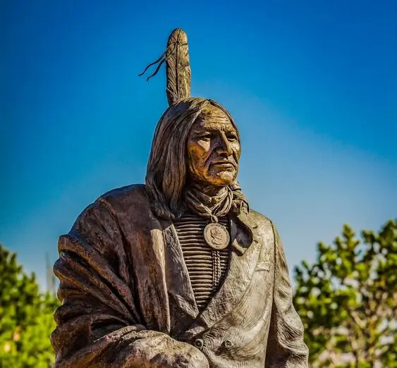 Bronze statue of a Native American man wearing traditional clothing and a single feather in his hair against a clear blue sky.