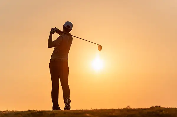 Silhouette of a golfer swinging a club at sunset on a grassy hill.