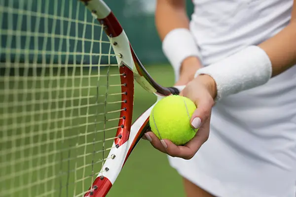 Close-up of a tennis player holding a yellow tennis ball near a red and white racket in front of a net.