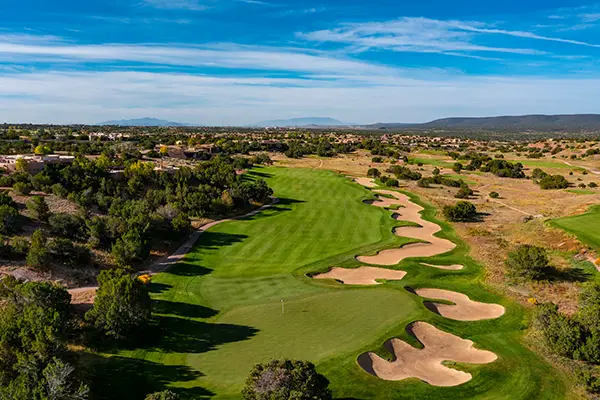 Aerial view of a golf course fairway with sand bunkers, surrounded by trees and residential houses under a blue sky.