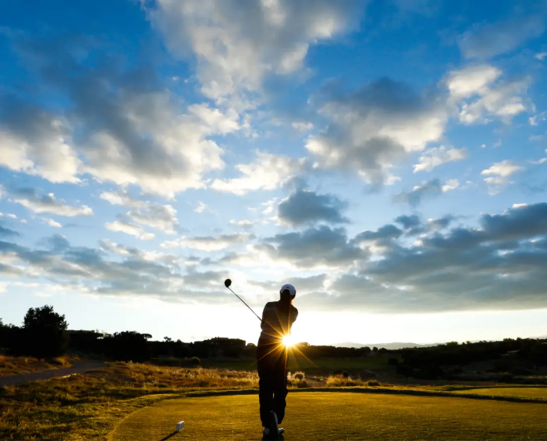 Golfer hitting a golf ball at sunset with a partly cloudy sky.