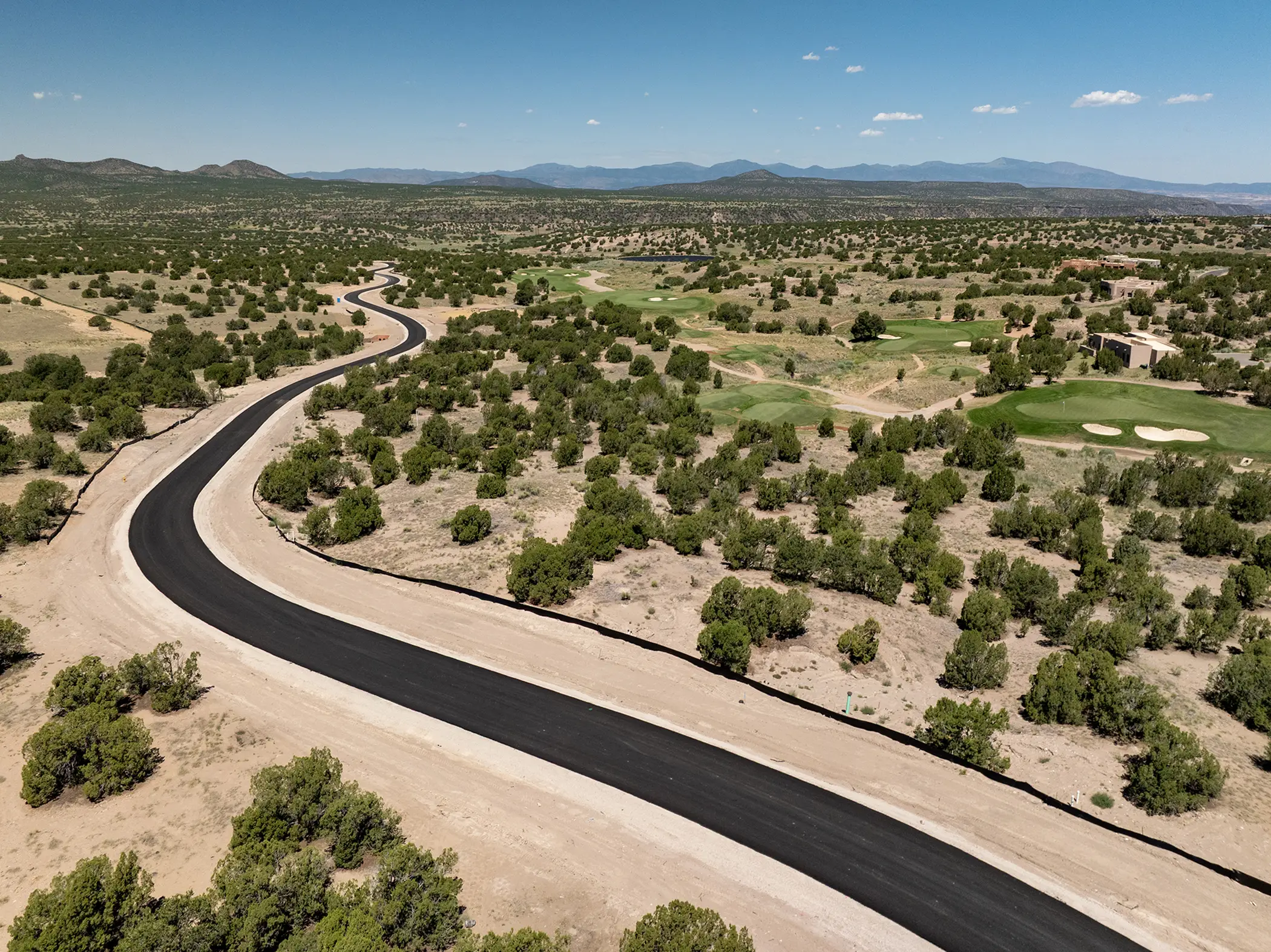A winding freshly paved road curves through a dry, shrub-covered landscape with a golf course and mountains in the distance under a clear blue sky.