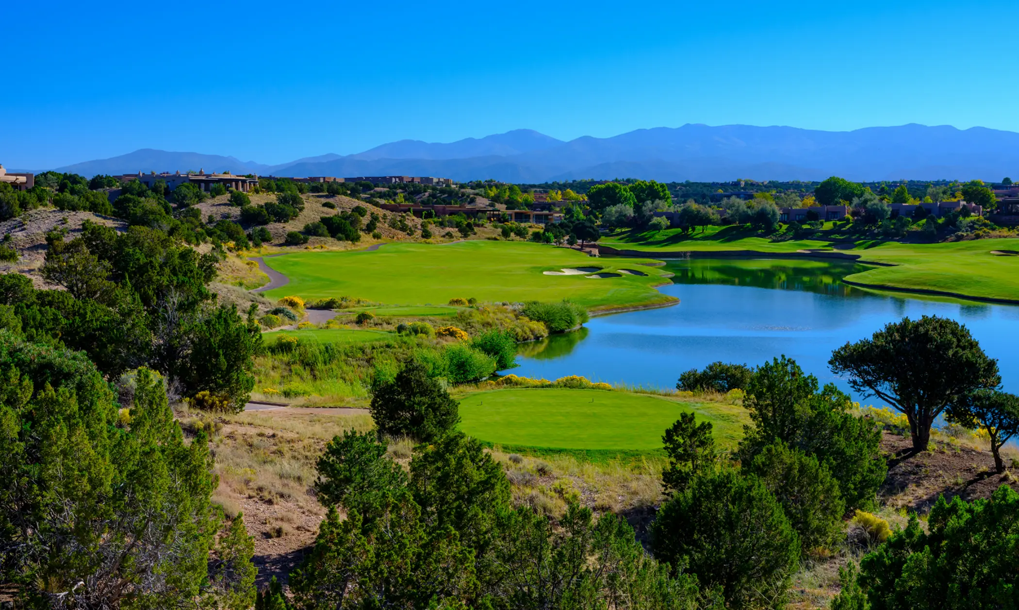Golf course with green fairways, a water hazard, surrounding trees, and mountains in the distance under a clear blue sky.