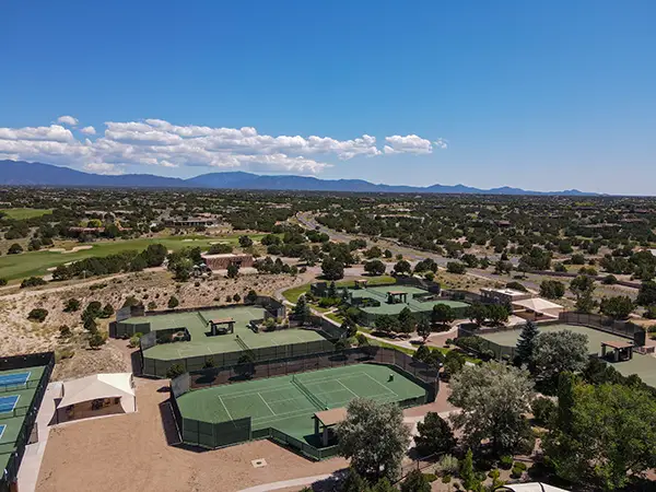 Outdoor tennis court with players engaged in a match, surrounded by greenery and buildings under a clear blue sky.