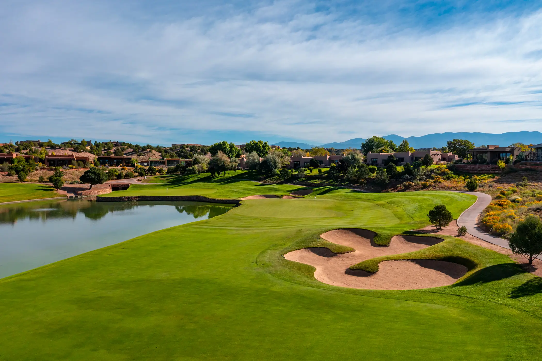Aerial view of a golf course with green fairways and sand paths, surrounded by desert terrain and scattered trees, mountains in the distance under a blue sky with clouds.