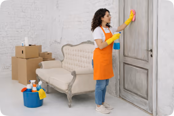 Woman in orange apron and yellow gloves cleaning a wooden door with a cloth and spray bottle in a room with a beige sofa and moving boxes.