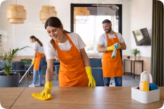 Three people wearing orange aprons and yellow gloves cleaning an office space with a woman wiping a wooden table in front.